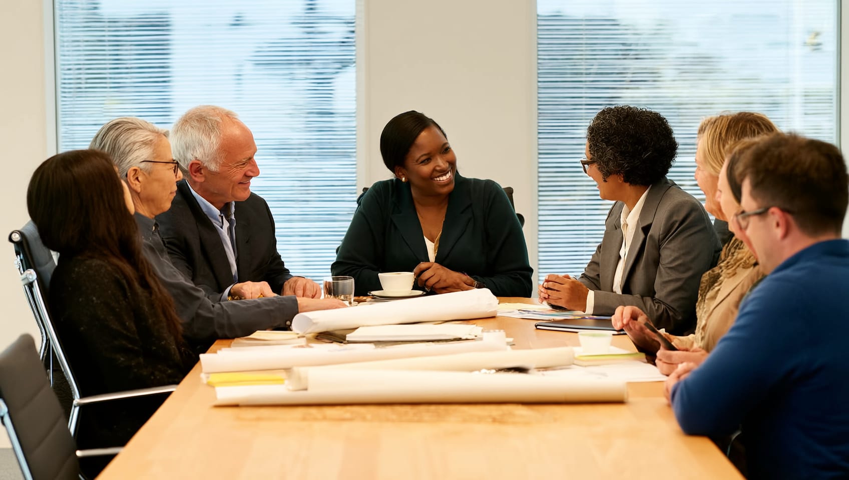 Company leaders of many backgrounds in warm conversation around a workshop table piled with plans