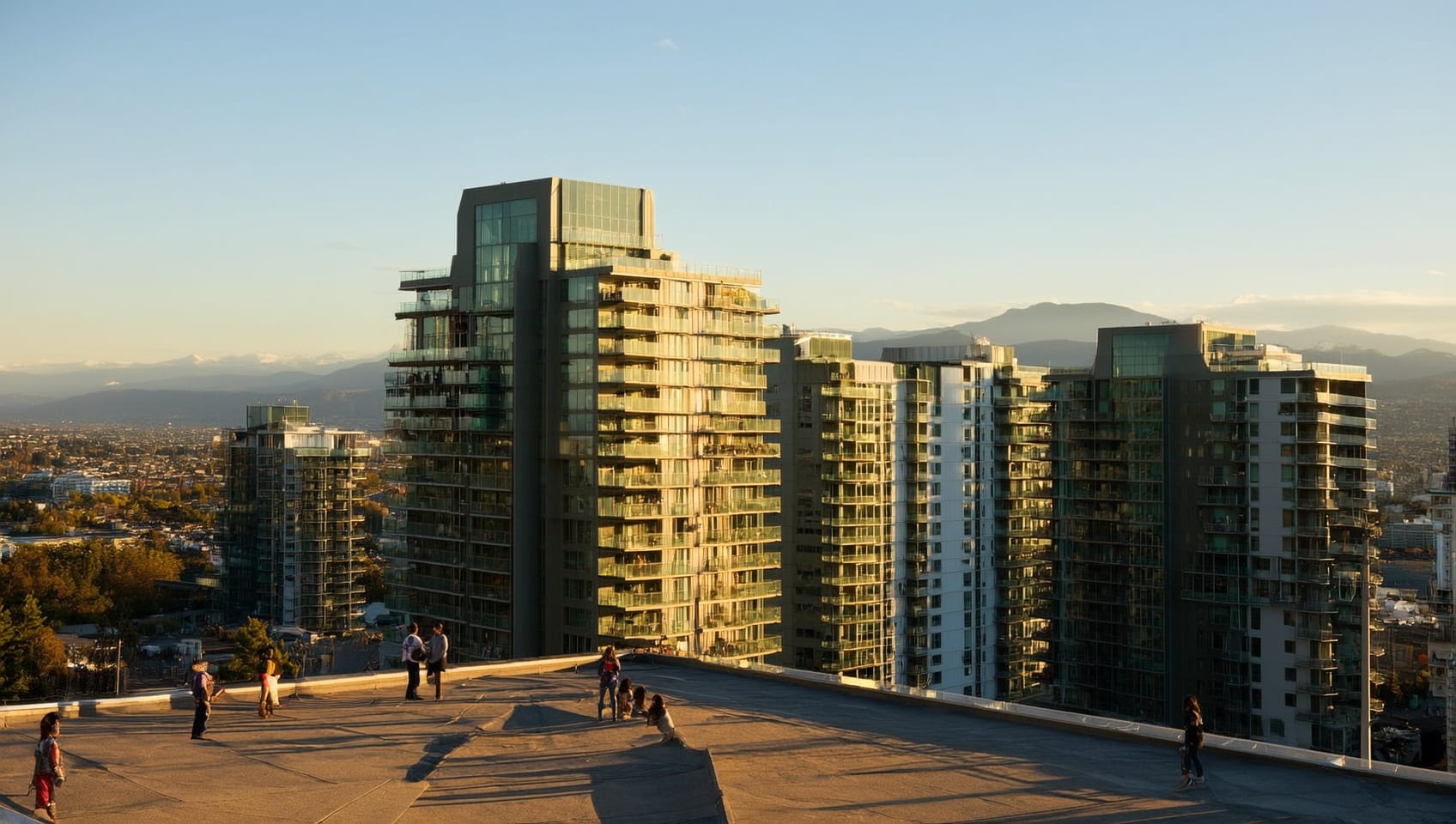 Sunlit residential glass towers with rooftop terraces rising over Vancouver toward distant mountains, people visible on a rooftop terrace